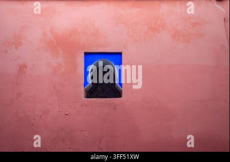 A small, ornate window is set into a pink wall, providing a glimpse of blue beyond the intricate latticework. Stock Photo