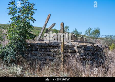 a weathered stack of old concrete fence posts sitting haphazardly in tall, dry grass behind a wire fence on a rural hillside under a clear blue sky. Stock Photo