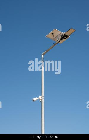 a tall, metal street light pole equipped with a small solar panel, an LED light fixture, and a security camera, standing against a vast, clear blue sk Stock Photo