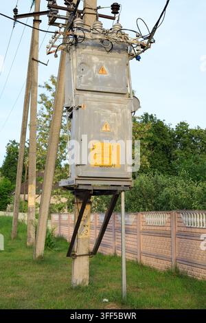 Old power transformer on a concrete pole near a fence. Rural electricity distribution infrastructure with high voltage cables stands against lush gree Stock Photo