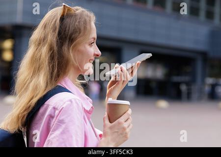 Smiling young woman with backpack and coffee uses voice assistant on smartphone outdoors in the city, illustrating travel navigation, communication an Stock Photo