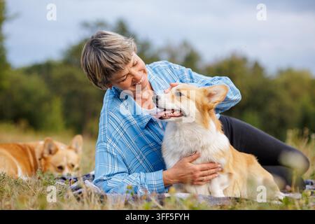Smiling mature woman lies on grass in a park hugging her corgi dog, portraying companionship, outdoor relaxation, and active pet friendly senior lifes Stock Photo