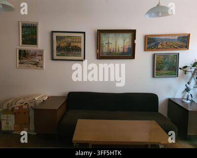 Interior view of a modest living room featuring a dark brown sofa centered against a white wall decorated with six framed paintings of various sizes Stock Photo