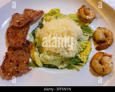 A Caesar salad served on a white plate, featuring fresh romaine lettuce leaves coated in creamy dressing, topped with a generous mound of shredded Stock Photo