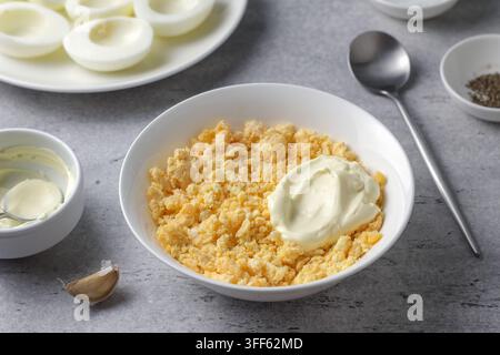 Crumbled hard boiled egg yolks in a bowl, garlic, mayonnaise and seasonings, prepared as filling for stuffed eggs on a gray textured surface Stock Photo