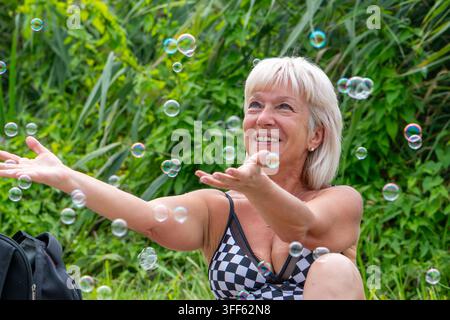 A smiling senior woman reaches toward floating soap bubbles in a green garden. Stock Photo