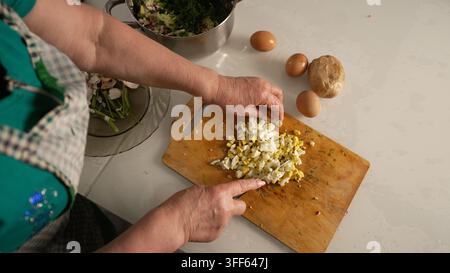 Overhead view of food prep with natural lighting, warm wood tones against white surface, focused hands slicing eggs, fresh ingredients arranged nearby Stock Photo