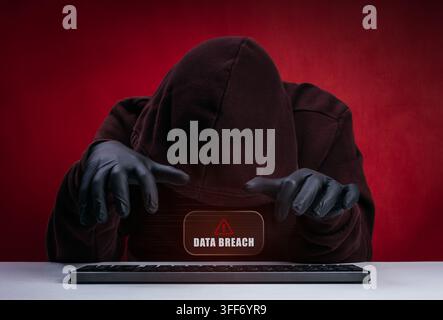 Hooded hacker wearing black gloves using keyboard computer during data breach. Cyberattack, unauthorized access, information theft, security threats, Stock Photo
