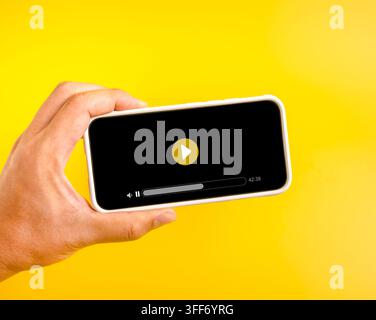 Hand holding smartphone with video player interface on vibrant yellow background, representing mobile streaming, online entertainment, digital media a Stock Photo