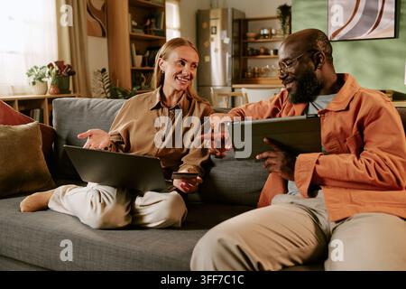 Caucasian middle aged woman and African American middle aged man sitting on sofa, shopping online using laptop, digital tablet and credit card. Ideal Stock Photo