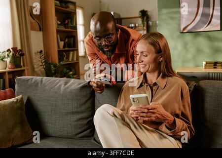 African American adult man showing credit card to smiling Caucasian adult woman using smartphone for online shopping. Scene suitable for Black Friday Stock Photo