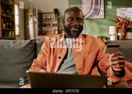 Middle aged African American man holding credit card using laptop for black friday shopping at home. Visual representing digital banking and ecommerce Stock Photo