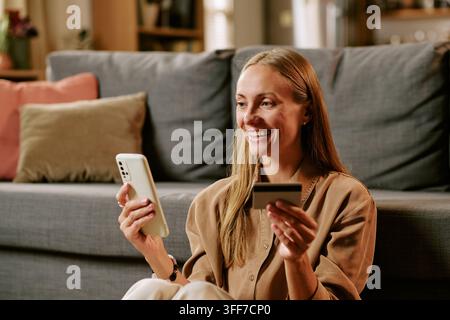 Smiling young adult Caucasian woman using smartphone and credit card for black friday shopping at home. Image suitable for e-commerce advertising and Stock Photo