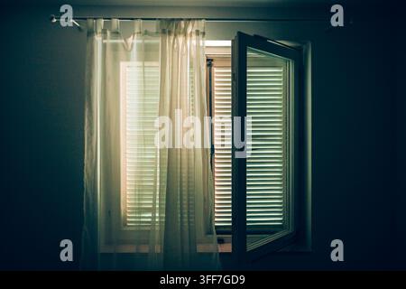 Interior shot from a dim room looking toward an open window with white curtains, where bright exterior daylight filters in through horizontal shutter Stock Photo