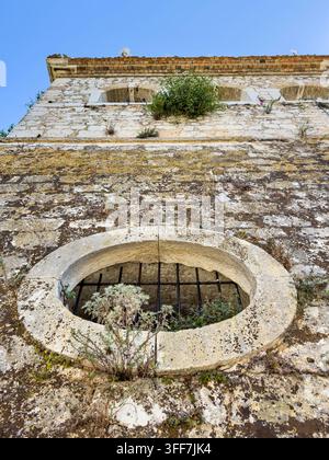 Low angle view of a weathered stone wall with a barred oval opening and wild plants at New Fortress in Corfu Town, Greece Stock Photo