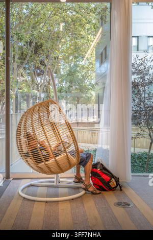 Barcelona - Spain - August, 25, 2026: Man Relaxing in Hanging Wicker Chair at Gabriel Garcia Marquez Library, Reading Nook by Large Window, Backpack Stock Photo