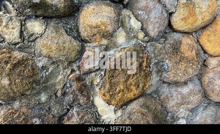 Close-up of a natural stone wall featuring tan and gray rocks with a rough textured masonry surface, suitable for architectural and background applica Stock Photo