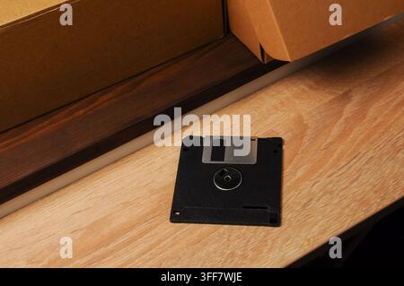 A single black 3.5 inch floppy disk is placed on a light wood grain surface, with a cardboard box partially visible behind it Stock Photo