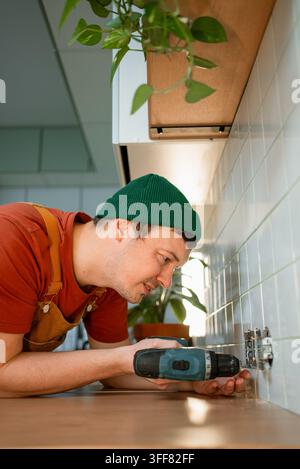 Man wearing an apron and green beanie actively installing an electrical socket on a tiled kitchen wall with a professional drill, demonstrating home renovation and diy skills Stock Photo
