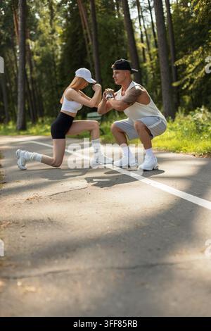 Fit young couple performing squats and lunges on a park path. Athletic man and woman doing leg exercises during an outdoor workout in a sunny summer p Stock Photo