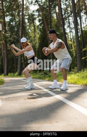 Athletic couple doing deep squats together during an outdoor fitness workout on a park path. Young fit man and woman exercising in stylish sportswear Stock Photo