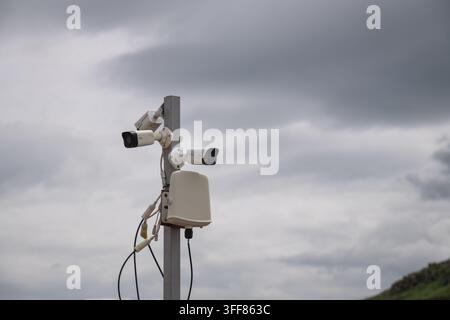 Multiple modern surveillance cameras mounted on a pole with exposed wiring, actively monitoring an outdoor area under a cloudy sky, representing conce Stock Photo