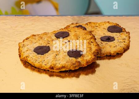 Close-up view of two large, freshly baked flat cookies topped with large chocolate discs, displayed on a shiny gold surface at a bakery or food stall. Stock Photo