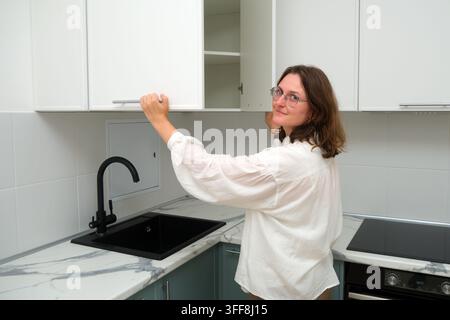 Woman opening white wall cabinet in new modern kitchen. Happy owner checking furniture after renovation or moving. Kitchen interior design and home im Stock Photo