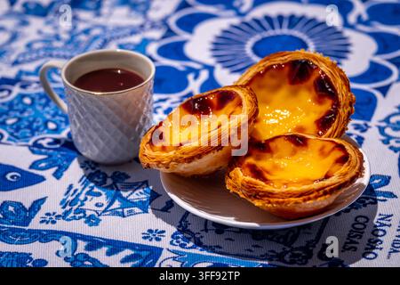 Three quarter shot of three pastel de nata custard tart on white plate and small porcelain coffee cup with geometric pattern, full of freshly-brewed espresso, on sunshine on blue and white tablecloth with traditional Portuguese azulejo tile motifs Stock Photo