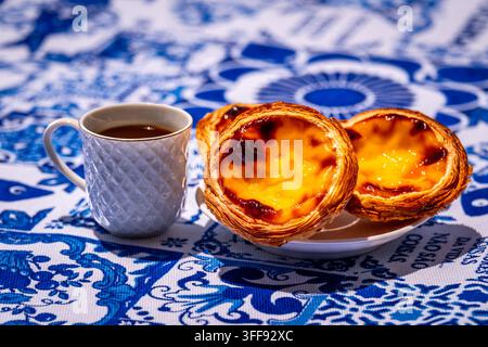 Three quarter shot of three pastel de nata custard tart on white plate and small porcelain coffee cup with geometric pattern, full of freshly-brewed espresso, in sunlight on blue and white tablecloth with traditional Portuguese azulejo tile motifs Stock Photo