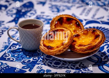 Three quarter shot of three pastel de nata custard tart on white plate and small porcelain coffee cup with geometric pattern, full of freshly-brewed espresso, outdoors on blue and white tablecloth with traditional Portuguese azulejo tile motifs Stock Photo