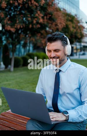A man is sitting on a bench in a green area, using a laptop and wearing headphones while smiling. Stock Photo