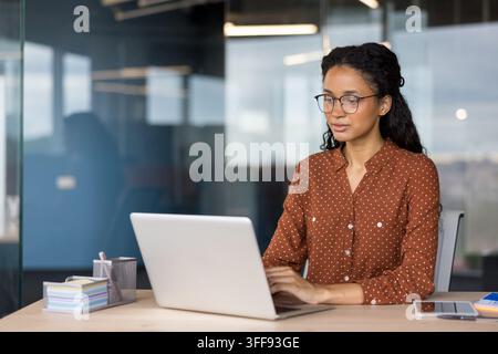 Young businesswoman wearing glasses and a dotted shirt, concentrating on her computer work, representing productivity and professional success in a contemporary corporate environment Stock Photo