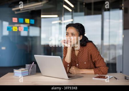 Young woman working in a modern office looking away from her laptop, experiencing lack of motivation, thinking, and feeling overwhelmed by work tasks and deadlines Stock Photo