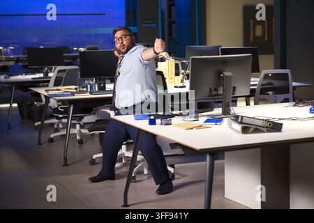 A playful medium shot of a plus-size businessman with glasses standing in a modern office at night. He mimics an archer pulling a bowstring, aiming at Stock Photo