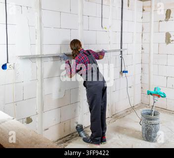 A female plasterer in overalls checks plastering guides on a wall made of aerated concrete blocks using a building rule. Stock Photo