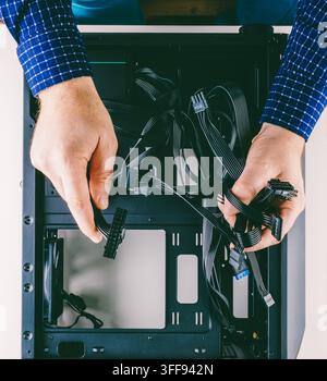 A man assembles a computer in his room at home, holding the wires from the power supply over the empty new black case lying on the table top view Stock Photo