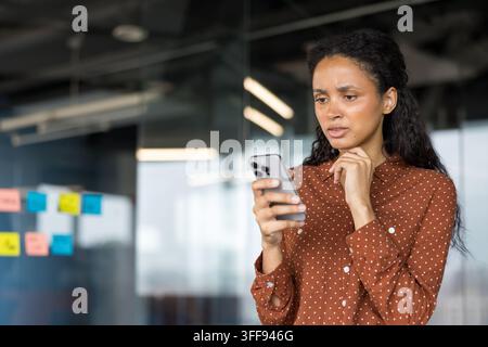 Young businesswoman in a modern office, focused and concerned as she reads her smartphone, illustrating digital communication, workplace stress, notifications and professional challenges Stock Photo