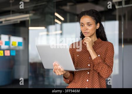 Young adult businesswoman analyzing data on laptop, contemplating new ideas for project development, standing in a contemporary workplace environment with glass walls Stock Photo