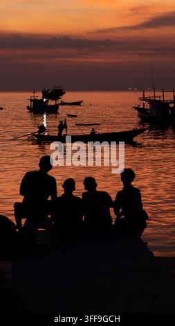 Four people sit as dark silhouettes on a rocky shoreline watching a vivid orange and red sunset over a calm bay filled with traditional wooden fishing Stock Photo