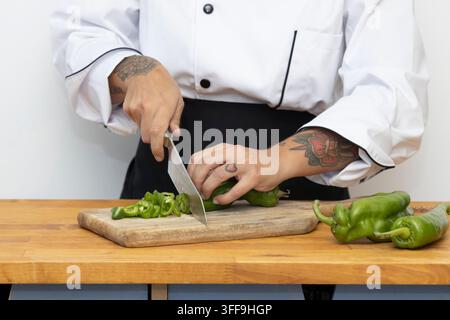A tattooed chef in a white professional jacket skillfully slices fresh green bell peppers on a wooden cutting board, with whole peppers arranged nearb Stock Photo