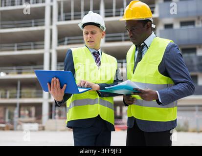 Two architects, Caucasian and African-american men in warnvests, standing on building site and discussing construction project. Stock Photo