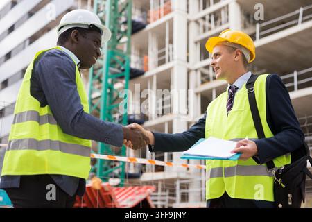 African-american and European engineers standing on construction site and shaking hands after concluding agreement. One man holding documents in hands Stock Photo