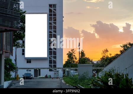 Sunset paints sky in warm hues. Large blank billboard stands tall. Building rises behind greenery. Vehicle rests near entrance. Ideal for advertising Stock Photo