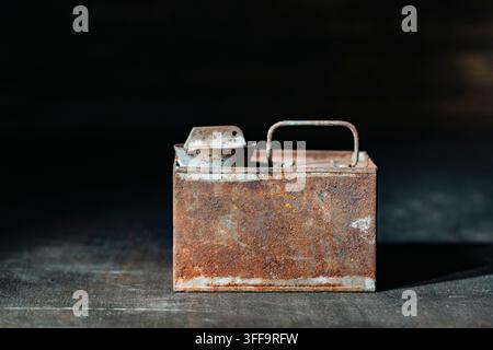 Vintage rusty metal canister. Old industrial rectangle tin can with handle covered in heavy rust and patina standing on scratched dark surface inside Stock Photo