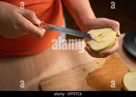 Close up of female hands peeling a fresh apple and removing its seeded core with a sharp knife on a cutting board in the kitchen. Vegan, diet, detox, Stock Photo
