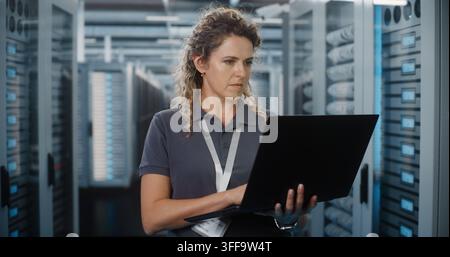 Focused Female Network Engineer Types on Laptop Computer While Standing Near Server Cabinets in Data Center. Hardware Configuration and Server Diagnos Stock Photo