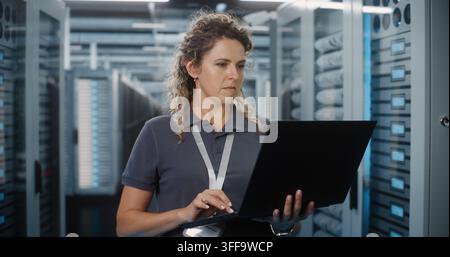 Focused Female Network Engineer Types on Laptop Computer While Standing Near Server Cabinets in Data Center. Hardware Configuration and Server Diagnostics Ensure Continuous Cloud Connectivity. Stock Photo