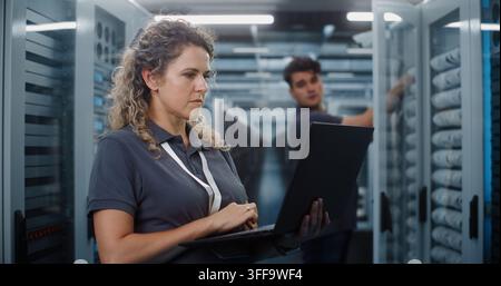 In Modern Data Center Server Room, Female IT Specialist Works on Laptop While Male Colleague Inspects Server Racks. System Administration and Teamwork Ensure Continuous Cloud Uptime. Big Data Concept. Stock Photo