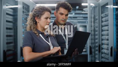 In Modern Data Center Server Room, Female IT Specialist Works on Laptop While Male Colleague Inspects Server Racks. System Administration and Teamwork Ensure Continuous Cloud Uptime. Big Data Concept. Stock Photo